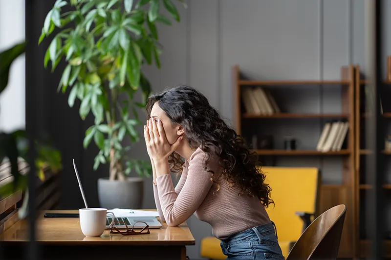 a tired woman covers her face with her hands and struggles to work at home